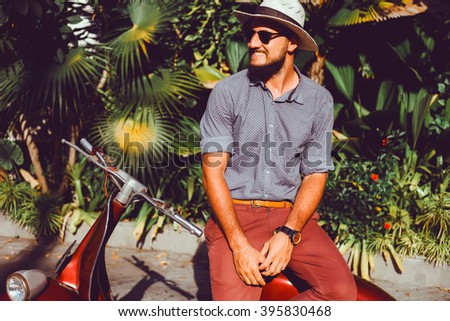 handsome in a light jacket, hat and red trousers, sunglasses with mustache and beard about posing retro scooter, fashionable clothes, brutal man, stylish outfit, Red, walk down the street