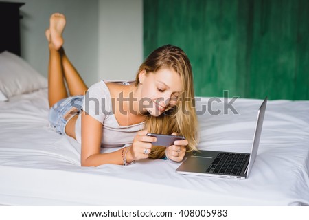 young blonde girl in t-shirt and denim shorts, hipster, lying on the bed at home in my room playing video games on your smartphone, writing letters, instagram, facebook, cell phone, close the laptop
