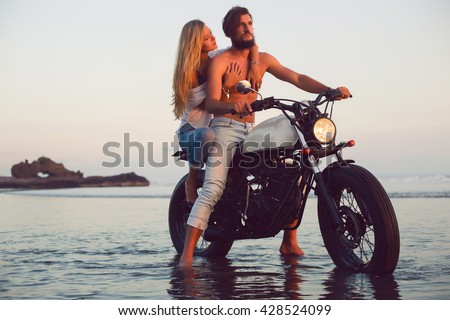young beautiful couple hipsters in stylish clothing for a retro motorcycle on the street, outdoor portrait, posing, in jeans and t-shirts, bearded guy, blonde girl, travel together, ocean, beach