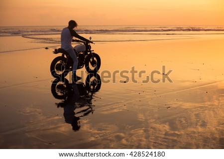 young man posing in sunset, on vintage motorcycle, near ocean and beach, hipster style