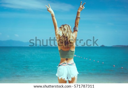 pretty woman posing in the sea, blue sky, hair wild, victory hand up!, outdoor portrait hipster, fashion model, pretty female, denim shorts, hippie, tattoo