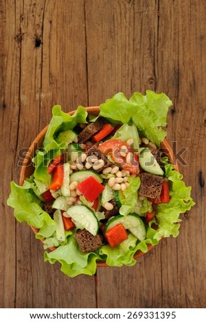 Vegetable salad with white beans, rye toasts, tomatoes, cucumber and lettuce in round bowl on wooden background top view with space