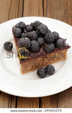Piece of tasty chocolate pie with ganache, decorated with fresh blackberries, lemon peel and icing sugar in white plate on wooden table