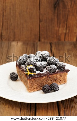 Piece of delicious chocolate pie with ganache, fresh blackberries and lemon peel decorated with icing sugar in white plate on wooden table
