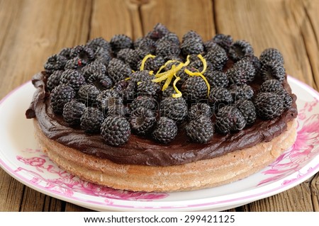 Tasty round homemade chocolate pie with ganache, decorated with fresh blackberries, lemon peel and icing sugar in white plate on wooden table