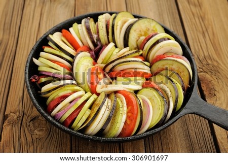 Raw vegetables layed for ratatouille made of eggplants, squash, tomatoes and onions in black cast iron pan on wooden table horizontal
