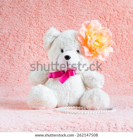 White teddy bear toy with orange flower barrette on head sitting on pink terry background