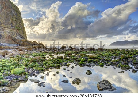 Haystack Rock with evening back lighting at low tide, Cannon Beach, Oregon. A large tide pool is in foreground with partly cloudy sky reflection. HDR fusion image. Warning sign on rock edited out..