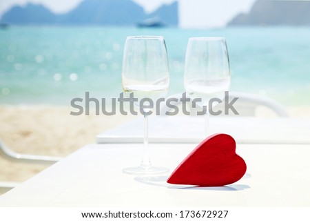 Red Heart On The Table With Two Glasses Of Wine Or Champagne And Sand And The Sea On The Background.