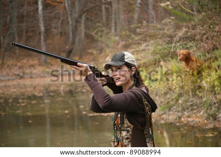 Woman Duck Hunter wearing Cap and Camo Waders Shooting Rifle in Pond with Dog in Background