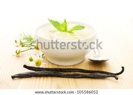 White bowl of vanilla yogurt with vanilla beans, flowers and a spoon on wood