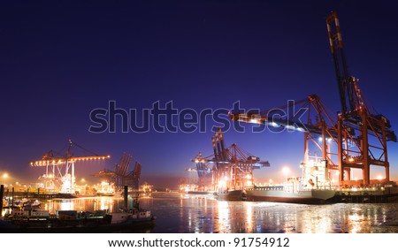 Panorama image of the illuminated cargo port in Hamburg at night with container terminals, cargo ships and cranes and a clear blue sky.