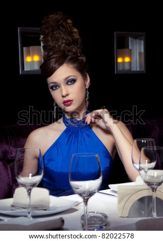 Very beautiful glamour girl in a dark blue dress sits at restaurant at a table