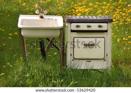 Old run-down kitchen sink (with tap and pipes, on a wooden stand) and kitchen stove (with three knobs, a closed oven, and a closed top), out on a field with grass and dandelions, sunny summer day