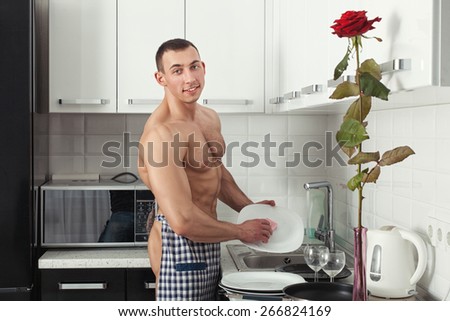 Bodybuilder man standing in the kitchen wearing an apron and washes the dishes.