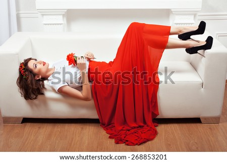 Girl lying on the sofa, her red dress and a flower in her hand. Interior and white sofa.