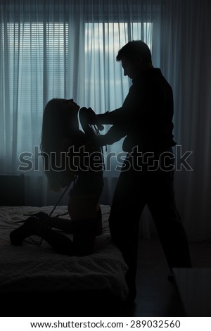 Only visible silhouettes of men and women. Man tyrant torturing a woman in a dark room on the bed.