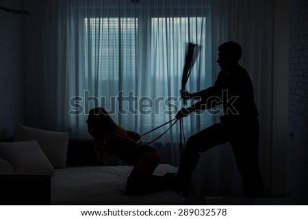 Black contours silhouette.  Man beats a woman with a whip in a dark room on the bed.