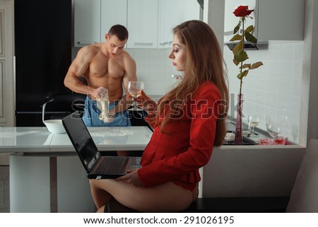 Foreground girl behind a laptop. She drinks wine. The man is out of focus cook. They are in the kitchen.