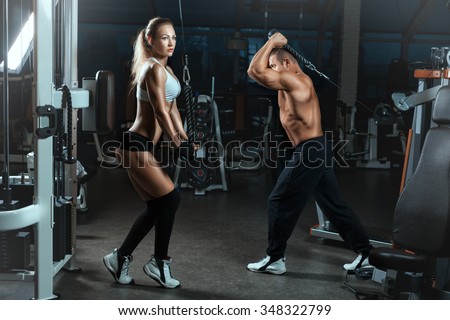 Girl and the man go in for sports in the hall with machines for bodybuilders. They lift heavy weights to build muscle.