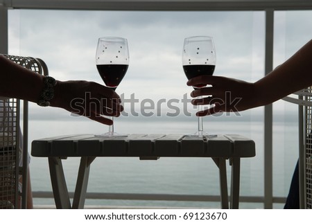 Hands of two people holding glasses of red wine on a balcony overlooking a lake.