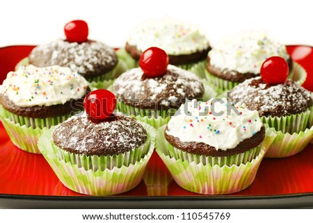 Delicious chocolate cupcakes topped with powdered sugar and maraschino cherry or with frosting and candies in red plate.