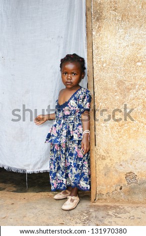 Cute but serious little black African girl in pink sunday dress next to her home door made of fabric; third world or developing country concept.