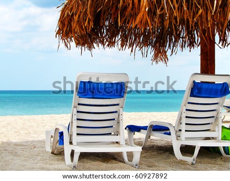 Two lounge chairs waiting for someone under a palapas at the beach of a tropical resort.