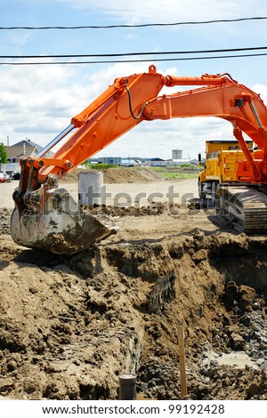 Construction: Bright orange mechanical digger removing dirt to install sewage and water line for a new neighborhood in rural North America with yellow dump truck behind it.