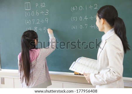 Female teacher standing next to the elementary Japanese girl who solves the problem of blackboard