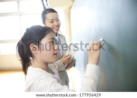 Female teacher standing next to the elementary Japanese girl who solves the problem of blackboard