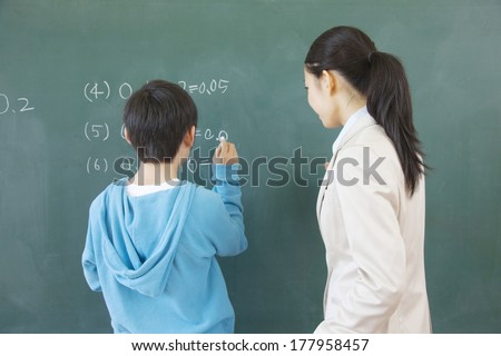 Female teacher standing next to the elementary Japanese boy who solves the problem of blackboard