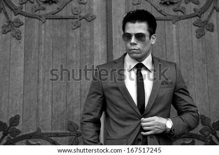 Businessman wearing sunglasses and gray suit and tie, in front near building door. Black & white photo