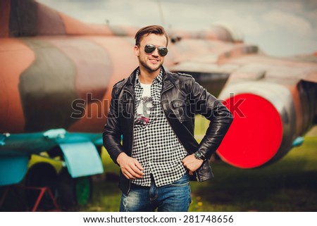 a man in a leather jacket and sunglasses smiling. planes in the background