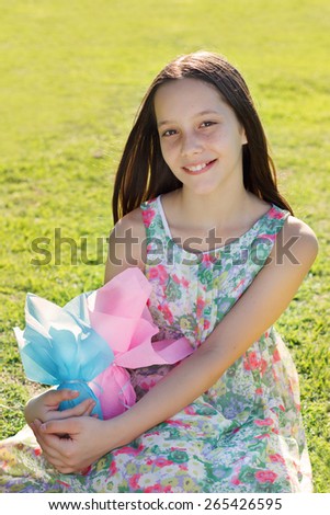 Beautiful smiling teen girl with freckles holding Easter chocolate eggs in colorful paper on green grass in park