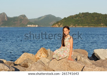 Happy smiling beautiful teen girl in dress with long brown hair sitting on stones on beach near sea. Selective focus. Toning effect
