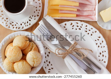 Brazilian snack pao de queijo (cheese bread) on white plate with butter, cheese, ham, cup of coffee on wooden table. Selective focus