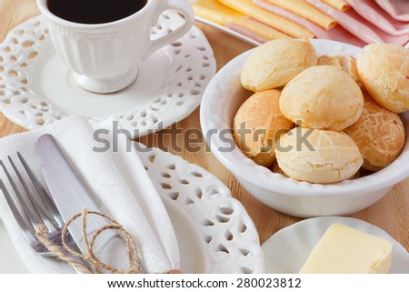 Brazilian snack pao de queijo (cheese bread) on white plate with butter, cheese, ham, cup of coffee on wooden table. Selective focus