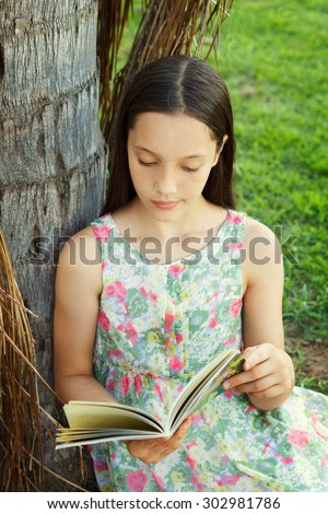 Cute teen girl reading book sitting on green grass near palm tree in park. Selective focus. Toning effect