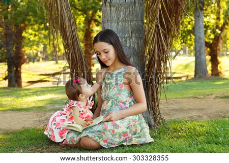 Cute sisters teen and baby girl reading book on green grass near palm tree in park. Selective focus