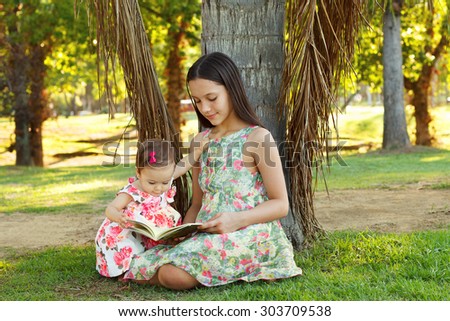 Cute sisters teen and baby girl reading book on green grass near palm tree in park. Selective focus
