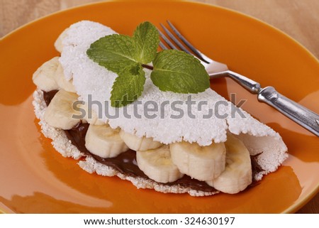 Casabe (bammy, beiju, bob, biju) - flatbread of cassava (tapioca) with banana and chocolate spread on orange plate on wooden table. Selective focus