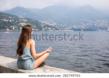 Attractive girl sitting by the lake in meditation pose, summer day, view from the back