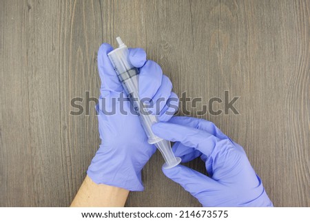 Man wear purple latex gloves and hold big plastic syringe on wood background.