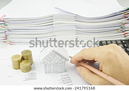 Man is holding pencil near house and gold coin on balance sheet with pile of paperwork as background.