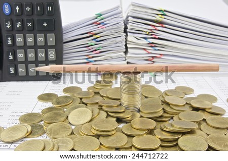 Brown pencil on stack silver and gold coins with pile of paperwork as background.