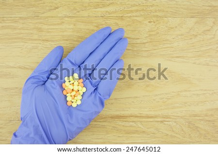 Yellow and orange pills in hand wear purple latex glove on wood background.
