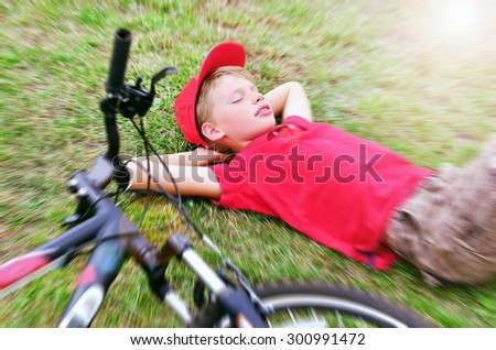 Boy in red T-shirt and cap is sleeping on the green grass with his bicycle with blur effect