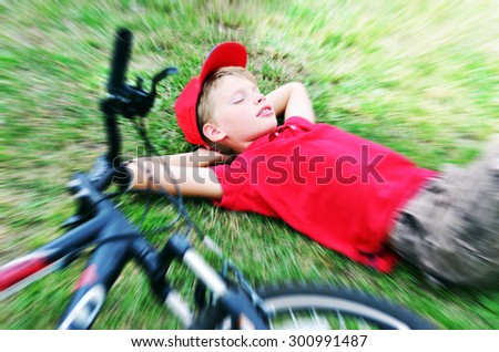 Boy in red T-shirt and cap is sleeping on the green grass with his bicycle with blur effect