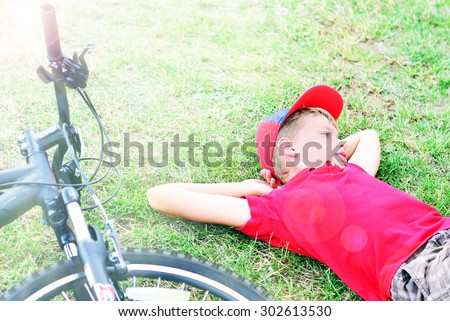 Boy in red T-shirt and cap is relaxing on the green grass next to his bicycle.